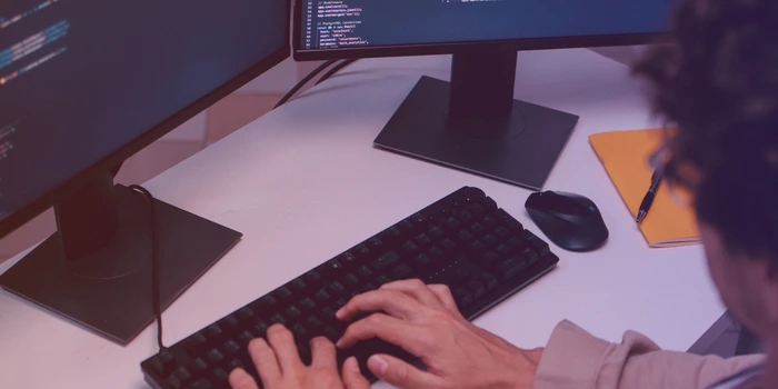 Hands typing on a keyboard with code on the monitor behind
