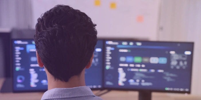 A developer seen from behind working across two monitors showing dashboards